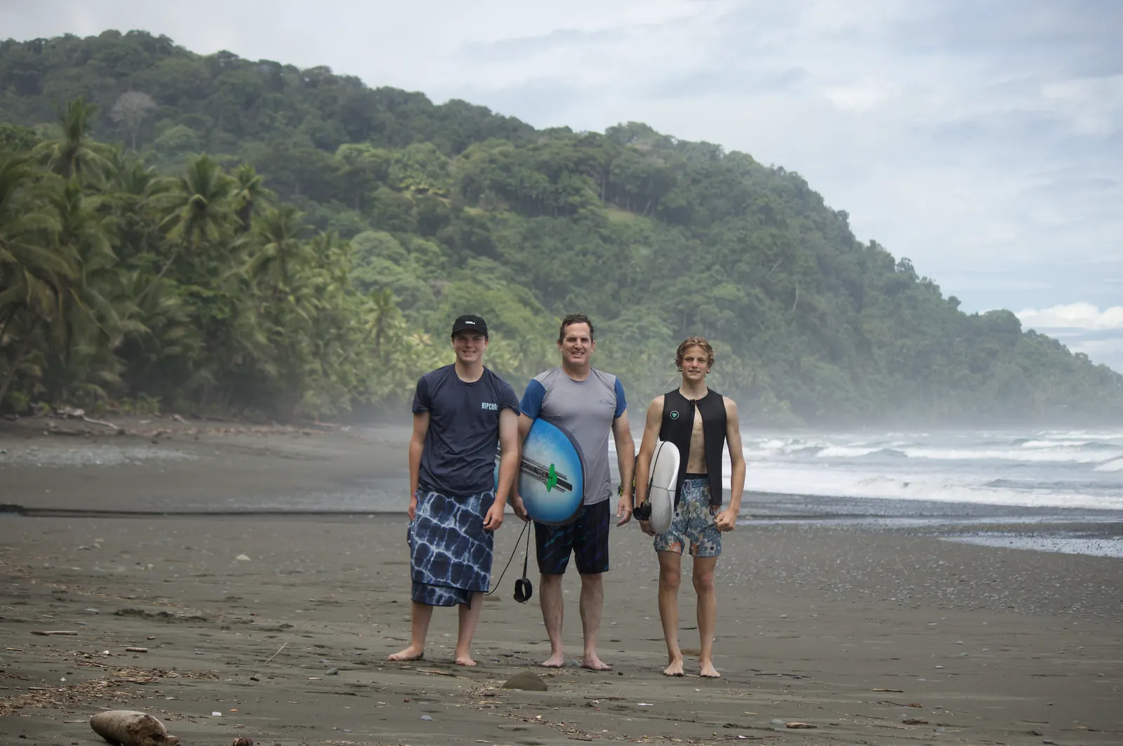 Three surfers on the beach with the jungle behind them
